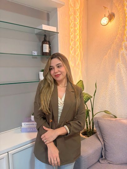 Dra. Alessandra de pé na sala, ao lado da estante de vidro com o frasco e a planta; atrás, a parede dourada e a arandela acesa.